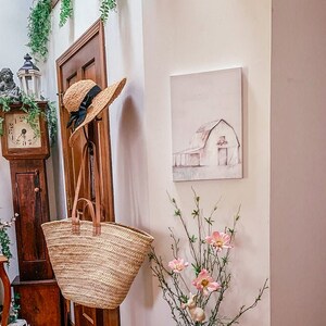 Puede incluir: Un sombrero de paja con una cinta negra cuelga de un gancho junto a una bolsa de mimbre. Un cuadro de una granja blanca con un techo rojo cuelga de la pared. Un ramo de flores rosas se encuentra sobre una mesa en la esquina.