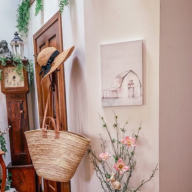 Puede incluir: Un sombrero de paja con una cinta negra cuelga de un gancho junto a una bolsa de mimbre. Un cuadro de una granja blanca con un techo rojo cuelga de la pared. Un ramo de flores rosas se encuentra sobre una mesa en la esquina.