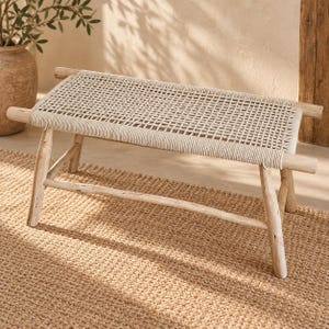 Puede incluir: Un banco rectangular con estructura de madera clara, cuatro patas inclinadas y un asiento trenzado de cuerda color blanco roto. El asiento presenta un patrón de rejilla y la estructura de madera incluye barras de soporte horizontales.