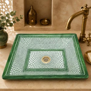 May include: A square ceramic vessel sink featuring a hand-painted green and white pattern of repeating scalloped shapes. The basin has a wide green border and a central circular drain with a metallic finish, set against a neutral stone countertop.