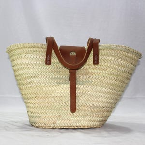 K&ouml;nnte beinhalten: Eine gro&szlig;e, geflochtene Strohtasche mit braunen Ledergriffen und einem Lederriemenverschluss. Die Tasche hat eine breite, offene Oberseite und einen abgerundeten Boden.