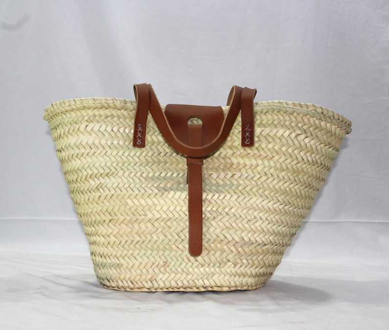 K&ouml;nnte beinhalten: Eine gro&szlig;e, geflochtene Strohtasche mit braunen Ledergriffen und einem Lederriemenverschluss. Die Tasche hat eine breite, offene Oberseite und einen abgerundeten Boden.