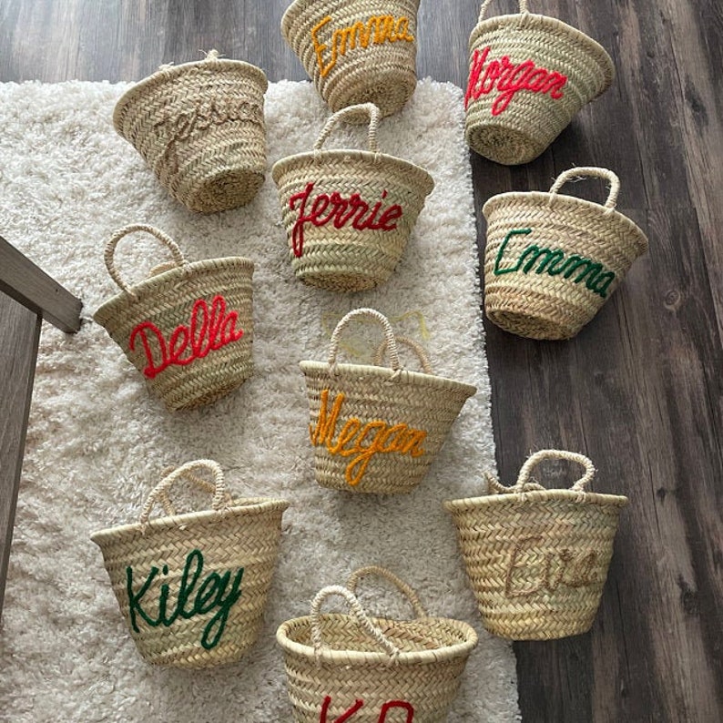 May include: Nine straw baskets with handles, each with a different name embroidered in colorful yarn. The names are: Emma, Megan, Ferrie, Della, Kiley, Emma, Morgan, and two unreadable names.