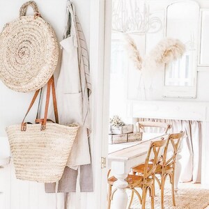 Puede incluir: Dos bolsos de mimbre colgando de una pared blanca con una puerta blanca en el fondo. Los bolsos est&aacute;n hechos de mimbre tejido natural y tienen asas de cuero. El bolso m&aacute;s grande es redondo y el m&aacute;s peque&ntilde;o es rectangular.