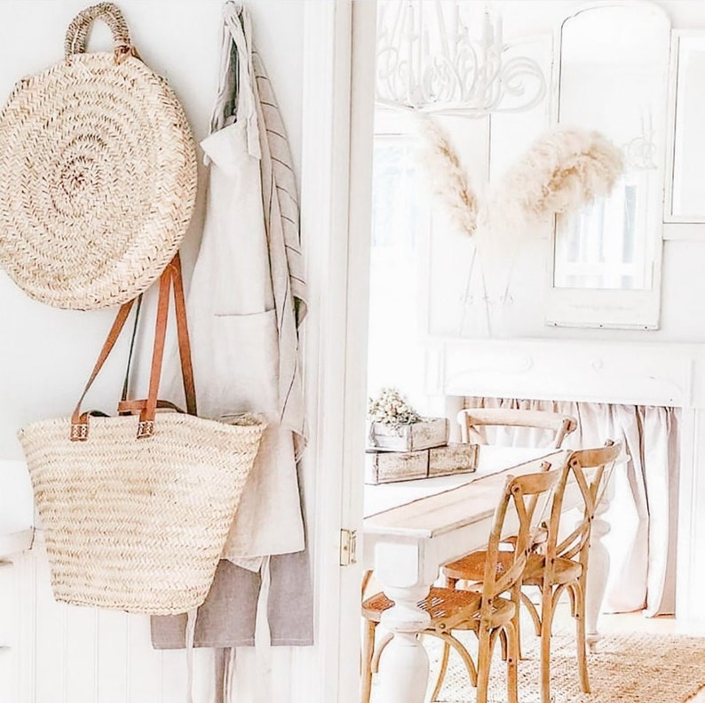 Puede incluir: Dos bolsos de mimbre colgando de una pared blanca con una puerta blanca en el fondo. Los bolsos est&aacute;n hechos de mimbre tejido natural y tienen asas de cuero. El bolso m&aacute;s grande es redondo y el m&aacute;s peque&ntilde;o es rectangular.