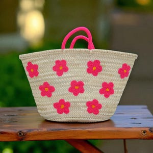 May include: A woven straw tote bag with bright pink flower appliqu&eacute;s and matching pink handles. The bag is a natural beige color, with the flowers evenly spaced across the front and sides. The handles are a soft, rounded shape.