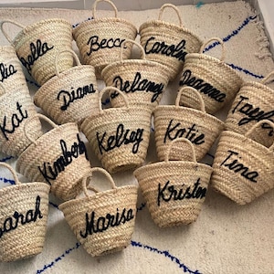 May include: A collection of 15 small straw tote bags with black embroidered names, including Julia, Bella, Kat, Sarah, Becca, Caroline, Diana, Delaney, Deanna, Taylor, Kimberly, Kelsey, Katie, Tina, and Kristen.