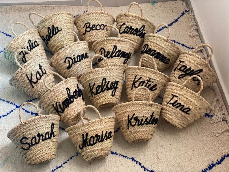 May include: A collection of 15 small straw tote bags with black embroidered names, including Julia, Bella, Kat, Sarah, Becca, Caroline, Diana, Delaney, Deanna, Taylor, Kimberly, Kelsey, Katie, Tina, and Kristen.