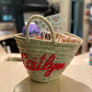 May include: A woven straw tote bag with a red embroidered name "Caitlin" on the front. The bag is filled with gifts, including a pink shirt with gold glitter lettering, a glittery tumbler, and a card that says "Legal to Drink Birthday Girl".