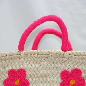 May include: Woven straw tote bag with bright pink handles and embroidered pink flower accents. The bag features a natural beige color with a textured weave, perfect for carrying items. The handles are made of a thick, soft material.