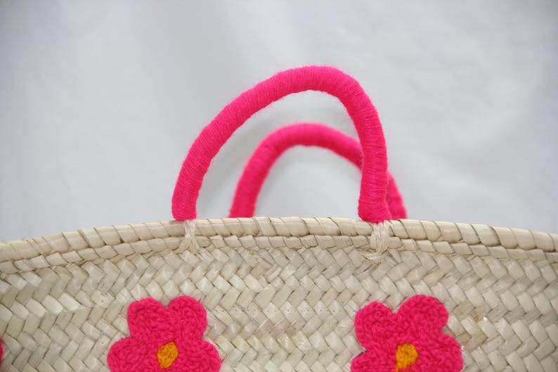 May include: Woven straw tote bag with bright pink handles and embroidered pink flower accents. The bag features a natural beige color with a textured weave, perfect for carrying items. The handles are made of a thick, soft material.