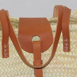 May include: Close-up of a woven straw tote bag with brown leather straps and a leather flap with a hole in the center. The straps are stitched with white thread.