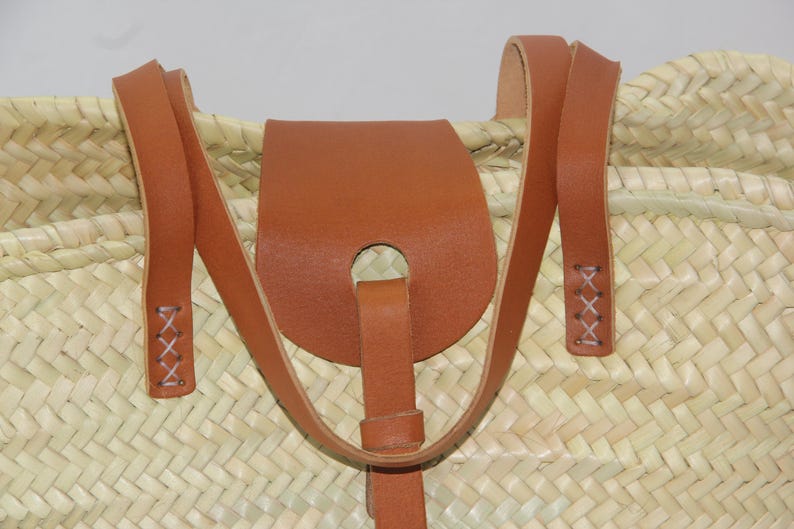May include: Close-up of a woven straw tote bag with brown leather straps and a leather flap with a hole in the center. The straps are stitched with white thread.