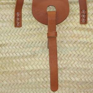 May include: A close-up of a woven straw bag with a brown leather strap and handle. The strap has a decorative loop with a stitched X pattern.