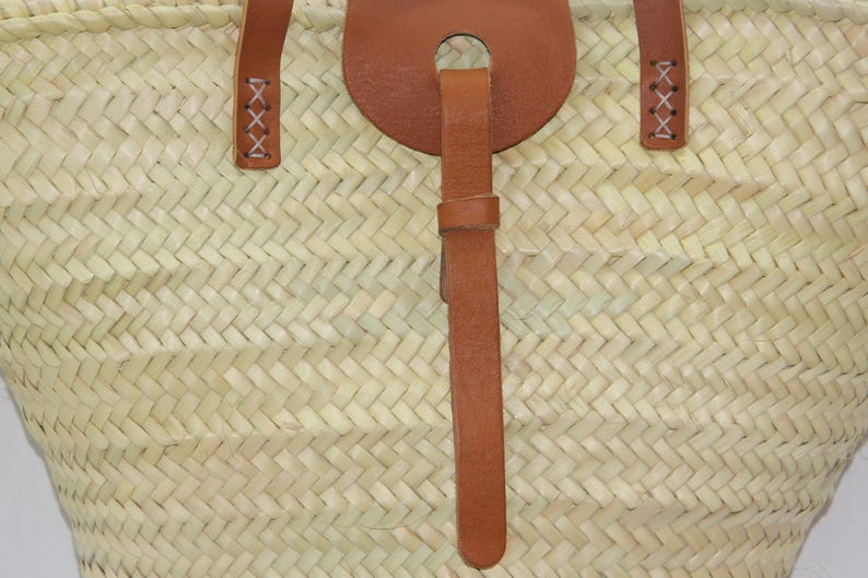 May include: A close-up of a woven straw bag with a brown leather strap and handle. The strap has a decorative loop with a stitched X pattern.