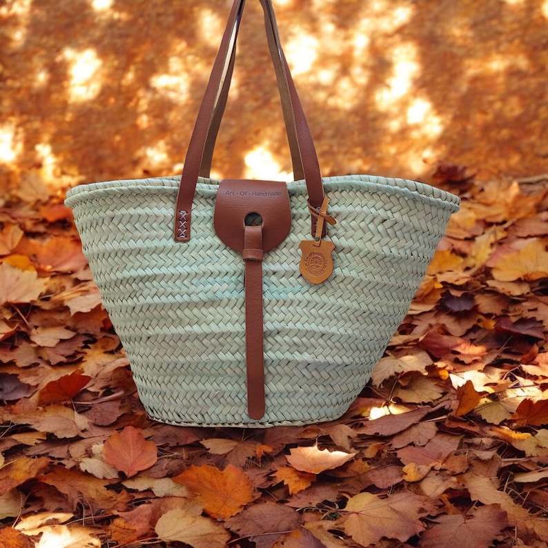 May include: A light blue woven tote bag with brown leather handles and a leather strap. The bag is sitting on a bed of fallen autumn leaves.