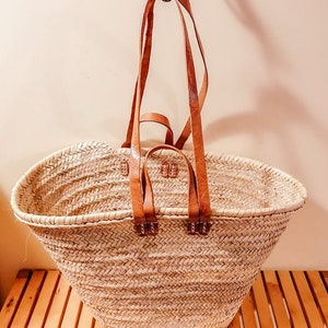 Puede incluir: Un bolso tote de paja grande, de forma ovalada, con asas de cuero marr&oacute;n. El bolso est&aacute; hecho de paja natural tejida y tiene un color beige claro.