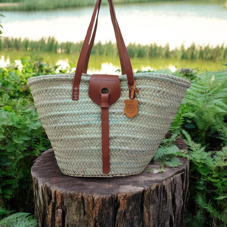 May include: A light green woven tote bag with brown leather handles and a leather strap. The bag has a leather tag with the text "Art of Handmade" on it. The bag is sitting on a tree stump.