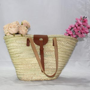 May include: A large, woven straw tote bag with brown leather handles and a leather strap. The bag is filled with pink and white flowers.