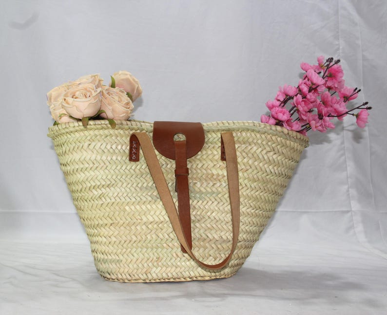 May include: A large, woven straw tote bag with brown leather handles and a leather strap. The bag is filled with pink and white flowers.