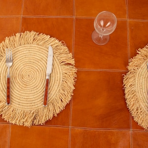May include: Two round woven placemats with fringed edges, each with a silver fork and knife. The placemats are on a brown leather surface. A clear glass wine glass is on the surface.