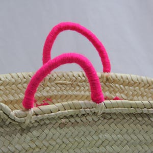 May include: Woven straw basket with bright pink handles. The basket is made of natural-colored straw and features two arched handles. The handles are wrapped in a vibrant pink material, adding a pop of color to the neutral tones of the basket.