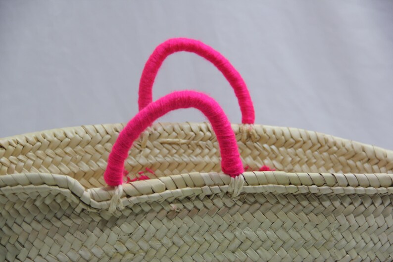 May include: Woven straw basket with bright pink handles. The basket is made of natural-colored straw and features two arched handles. The handles are wrapped in a vibrant pink material, adding a pop of color to the neutral tones of the basket.