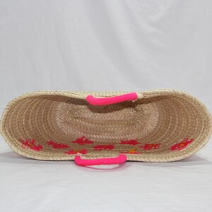 May include: Woven straw tote bag with bright pink handles and floral embroidery. The open-top basket has a natural beige color with pink accents. The handles are approximately 6 inches long.