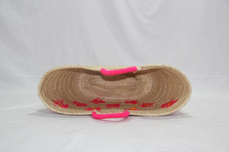 May include: Woven straw tote bag with bright pink handles and floral embroidery. The open-top basket has a natural beige color with pink accents. The handles are approximately 6 inches long.