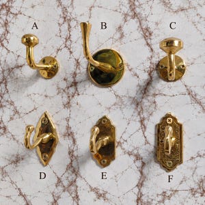 May include: Six gold-colored metal wall hooks labeled A through F. Hooks A, B, and C have circular mounting plates. Hooks D, E, and F have decorative, elongated mounting plates with visible screws. Each hook features a curved design for hanging.