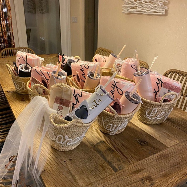 May include: A collection of small wicker baskets filled with pink and white towels, tumblers, and sunglasses. The baskets are decorated with the words "Beach", "Day", "Party", "Bride", and "Rita".