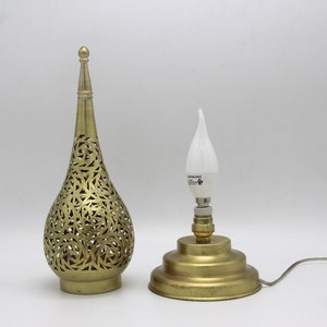 Op de afbeelding: Een messing tafellamp met een decoratieve, opengewerkte, traanvormige kap. De lamp heeft een eenvoudige, ronde voet en een heldere, traanvormige gloeilamp.