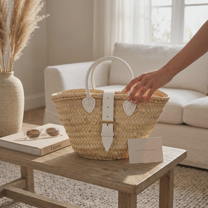 May include: A woven straw basket bag featuring white leather handles and a central white leather strap with a gold-toned buckle closure. The bag has a natural tan color and a textured, braided construction, providing a structured yet flexible shape.