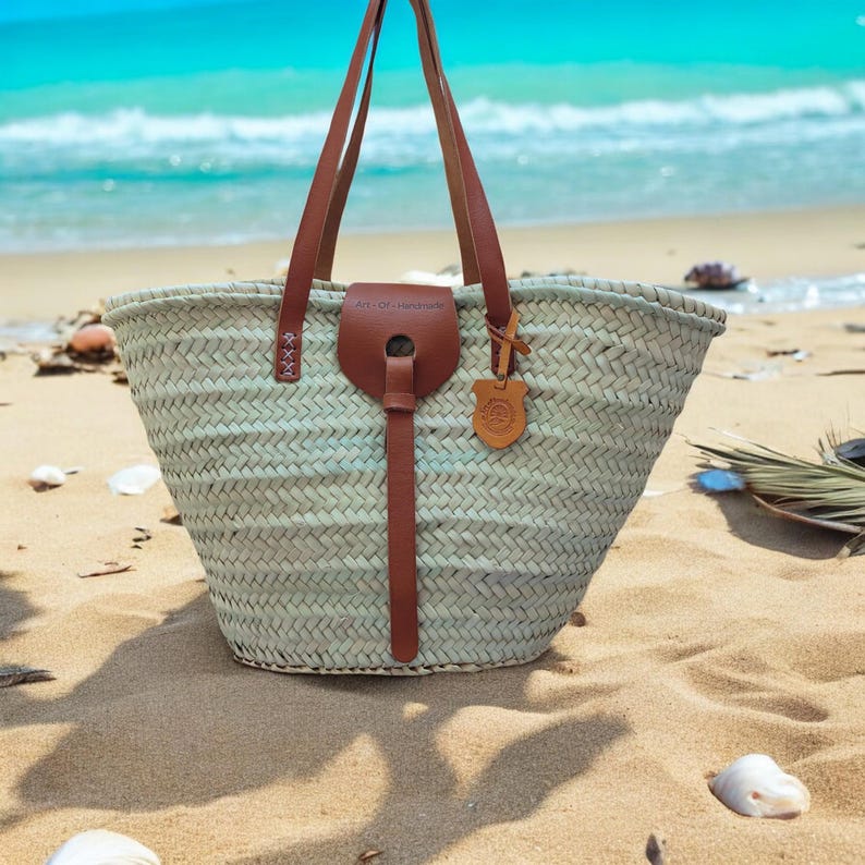 May include: A light blue woven tote bag with brown leather handles and a leather strap. The bag is sitting on a sandy beach with the ocean in the background. The bag has a label that says "Art of Handmade".
