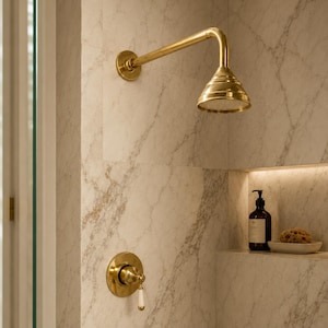May include: A wall-mounted brass shower head with a matching circular control valve featuring a white handle. The shower fixture has a polished metallic finish and is installed against a white marble wall with grey veining in a bathroom setting.