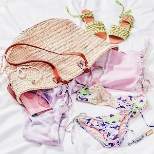 Puede incluir: Un bolso de paja con asa de cuero marr&oacute;n, una toalla a rayas rosa y blanca, un par de sandalias verdes y blancas, un bikini floral rosa y verde y una botella de perfume.