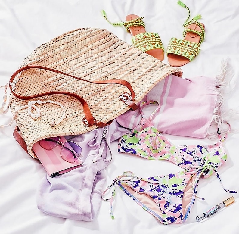 Puede incluir: Un bolso de paja con asa de cuero marr&oacute;n, una toalla a rayas rosa y blanca, un par de sandalias verdes y blancas, un bikini floral rosa y verde y una botella de perfume.