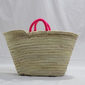 May include: A woven straw tote bag with bright pink handles. The bag has a wide, open top and a textured, basket-weave design. The handles are made of a soft, fuzzy material. The bag is suitable for carrying groceries or other items.