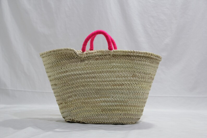 May include: A woven straw tote bag with bright pink handles. The bag has a wide, open top and a textured, basket-weave design. The handles are made of a soft, fuzzy material. The bag is suitable for carrying groceries or other items.