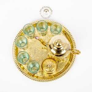 May include: A gold-colored Moroccan tea set on a round tray. The set includes a teapot, sugar bowl, and six small glasses. The tray and tea set have intricate embossed patterns. The glasses have a light green tint. The image includes the text "Art of handmade Moroccan crafts."
