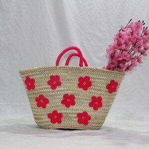May include: A woven straw tote bag with bright pink flower appliqu&eacute;s. The bag has pink handles and is filled with pink flowers. The bag is a light tan color and is sitting on a white surface.