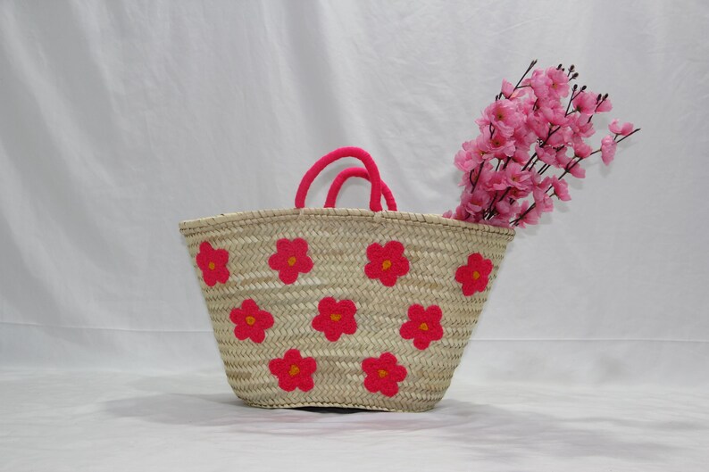 May include: A woven straw tote bag with bright pink flower appliqu&eacute;s. The bag has pink handles and is filled with pink flowers. The bag is a light tan color and is sitting on a white surface.