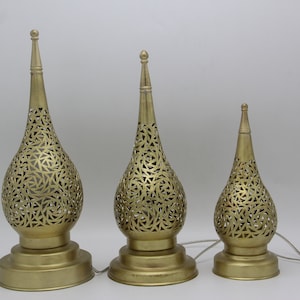 Op de afbeelding: Drie gouden metalen tafellampen met ingewikkelde uitgesneden ontwerpen in de vorm van tranen. De lampen zijn van verschillende grootte en worden van binnenuit verlicht.