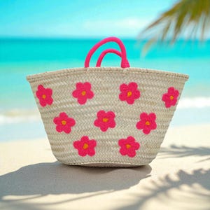 May include: A woven straw tote bag with bright pink flower appliqu&eacute;s and matching pink handles. The bag is on a sandy beach with a turquoise ocean in the background. A stylish and colorful beach bag.