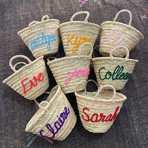 May include: Nine small straw baskets with colorful embroidered names, including "Sarah", "Claire", "Eve", "Colleen", "Jenna", "Kyan", "Caitlyn", and two others.