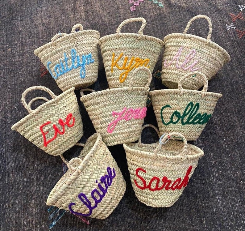 May include: Nine small straw baskets with colorful embroidered names, including "Sarah", "Claire", "Eve", "Colleen", "Jenna", "Kyan", "Caitlyn", and two others.