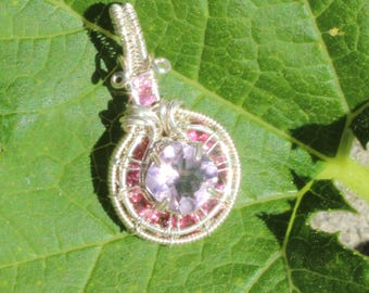 May include: A handcrafted pendant featuring a round, light purple gemstone encircled by smaller pink stones. The pendant is wire-wrapped in silver and rests on a large green leaf, creating a natural and elegant aesthetic. The pendant is a piece of jewelry.