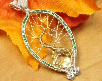 May include: A silver pendant with a wire tree design, featuring a yellow gemstone at the base and green accents around the oval frame. The pendant is set against a backdrop of fall foliage.
