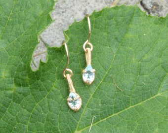 May include: A pair of gold-colored wire-wrapped earrings with teardrop-shaped, light blue gemstones. The earrings are displayed on a large, green leaf. The gemstones are set in a gold wire frame, and the earrings have a simple hook design.