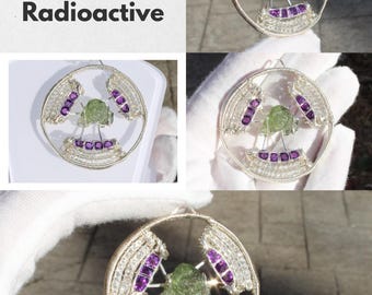 May include: A silver wire pendant featuring a central green gemstone, surrounded by purple amethyst stones and clear crystals. The word "Radioactive" is displayed in bold, black text, along with the words "Moldavite", "Amethyst", and "Rainbow Moonstone".